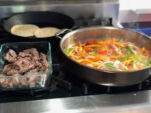 peppers and tortillas cooking on a stove, next to a container of meat